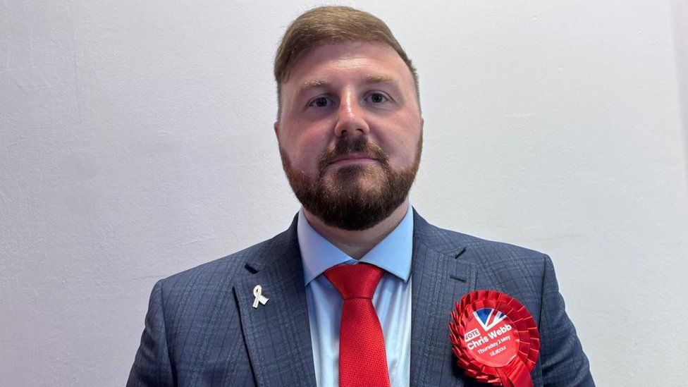 Chris Webb, a white man with brown hair and a beard looks into the camera. He's wearing a Labour rosette.