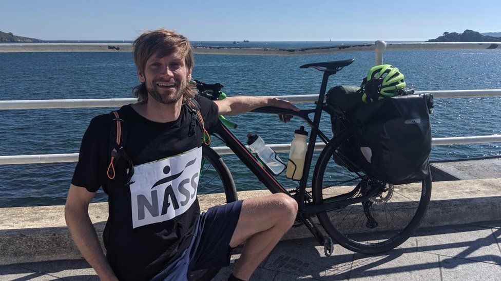Man next to a body of water with a bike, wearing a shirt that reads NASS