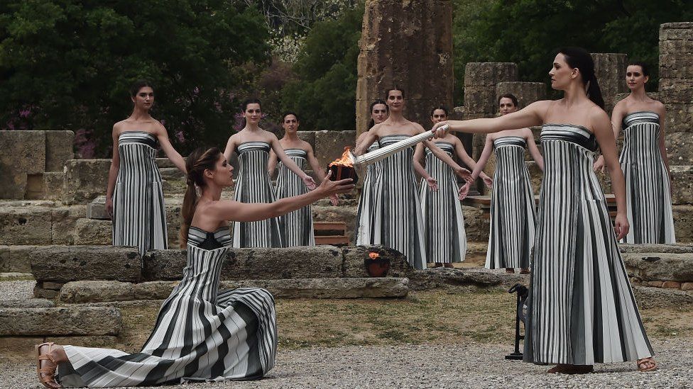 Woman lighting the Olympic torch from another woman who is on one knee. They are wearing black and white striped dresses