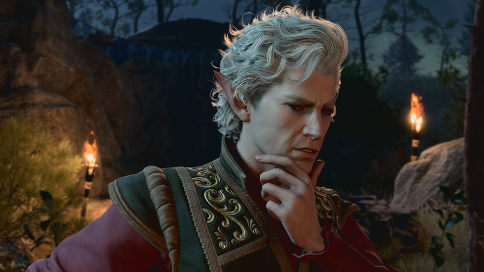 A man with white hair, pointed ears and unusually pale skin puts his hand to his chin, apparently deep in thought. He wears a high-necked, elaborate green jacket, with golden patterning on the shoulders. The Medieval garment has deep purple sleeves. He's in what appears to be a forested area. It's night-time, and two flaming torches attached to rocks in the background illuminate the scene