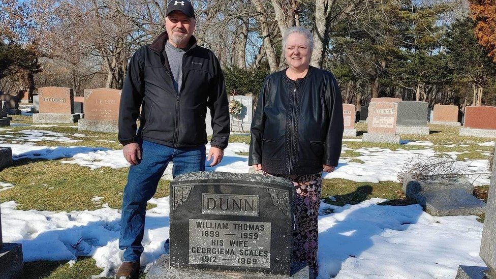 Man and woman stand next to grave stone as snow melts on ground