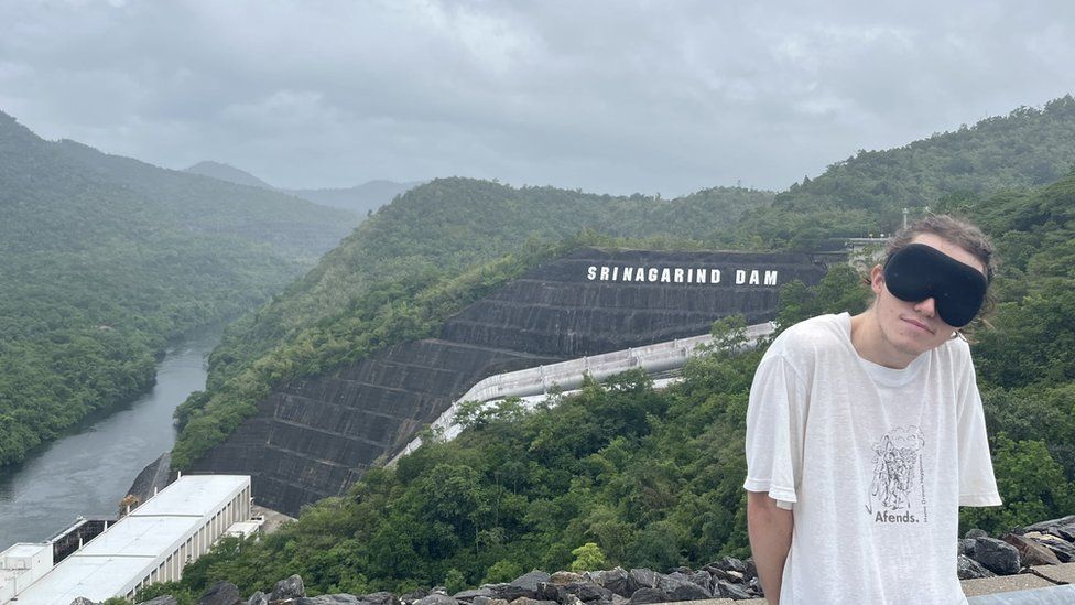 Ed Smith, blindfolded, standing beside view of dam in Thailand