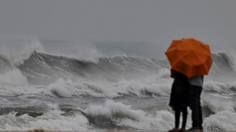 A couple stands holding an umbrella amid gusty winds as dark clouds loom over Marina beach in Chennai on December 8, 2022, ahead of Cyclone Mandous forecasted landfall in north Tamil Nadu-south Andhra Pradesh coasts. (Photo by Arun SANKAR / AFP) (Photo by ARUN SANKAR/AFP via Getty Images)