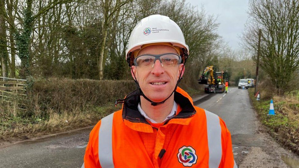 Man in hard hat and orange hi-viz stands on a road with the pothole machine in the background