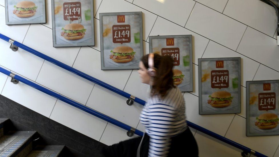 A commuter walks past food advertisement for a McDonalds burger, seen at Oxford Circus tube station in London, on 11th May 2018
