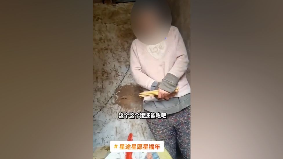 A screenshot of the video shows the woman with a chain around her neck standing in the hut