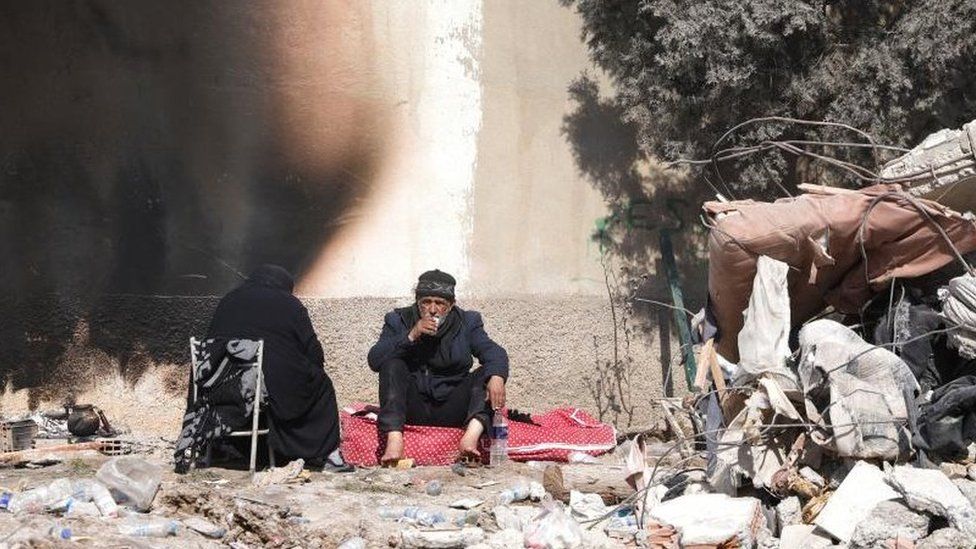 A man sits on a red cushion beside a ruined building