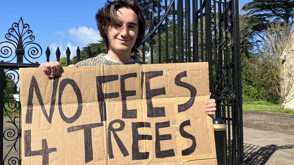 Henry Snowball holding a sign that says "No fees for trees" as he stands outside the iron gates of Cirencester Park