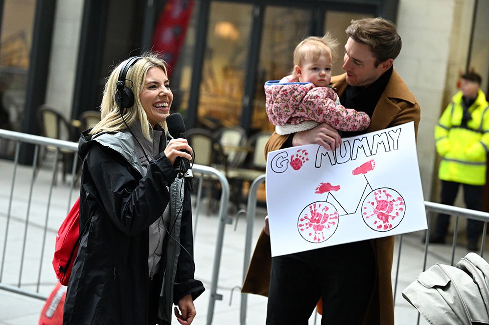 A woman holding a radio microphone and wearing large over-ear headphones smiles widely as she looks lovingly at a tall man with short brown hair who's holding a young child in his arms. The man is also holding a large white sign with "Go Mummy!" and a picture of a bicycle on it.