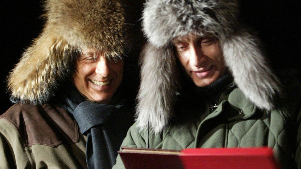 Russian President Vladimir Putin presents a book to then Italian President Silvio Berlusconi. Both wear large fur hats.