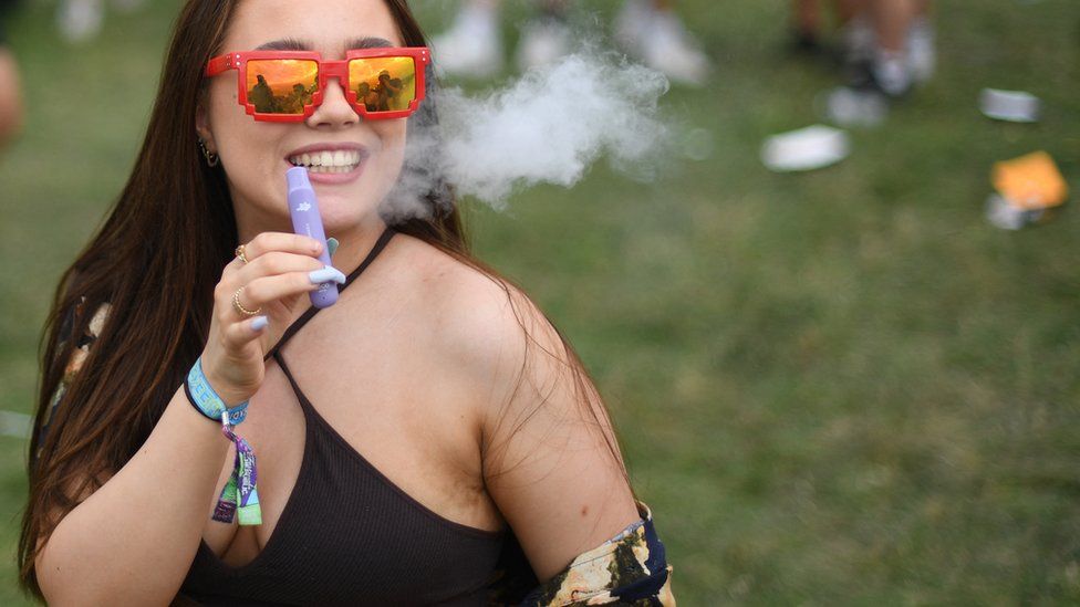 A young woman vapes at Reading festival. She is holding a lilac disposable vape, exhaling vapour. She is wearing a black bikini top and smiling