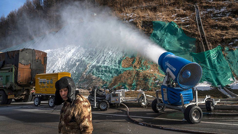 A workers stands next to a snow machine that is making artificial snow