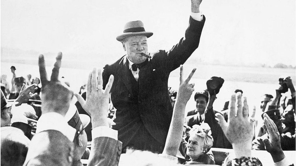 Winston Churchill, smoking a cigar, makes the V for Victory sign as he greets well-wishers from his car