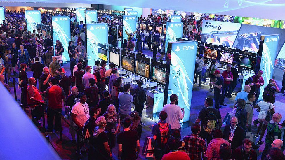 The showfloor of a gaming expo viewed from an elevated angle. It's very crowded, and there are rows of TV screens hooked up to consoles. People can be seen playing a range of different games. The scene is dotted with tall, brightly light boards displaying the PS3 logo