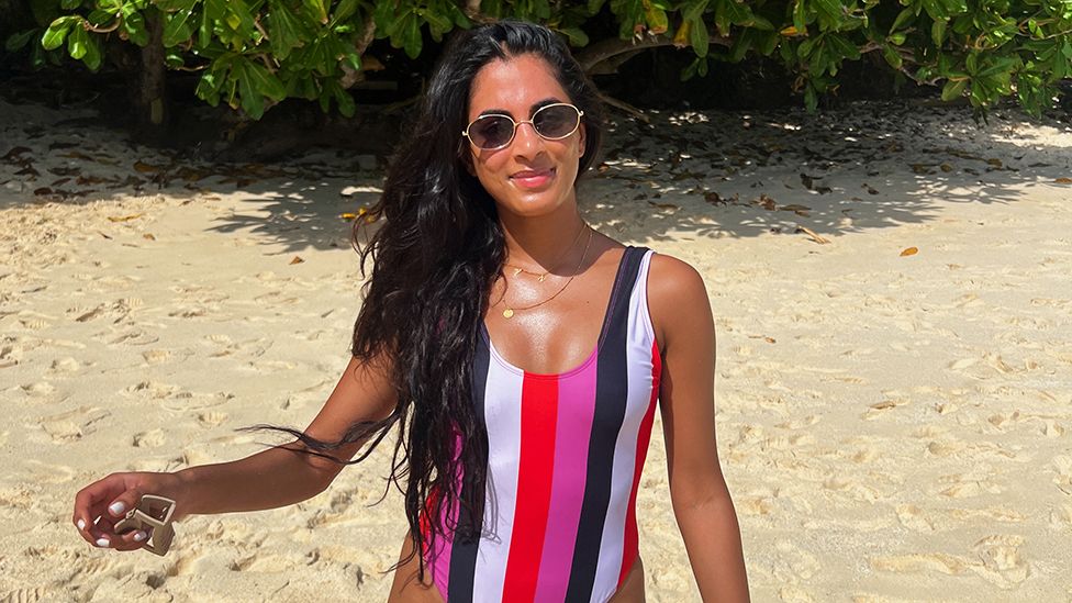 Tanvi is on the beach, wearing sunglasses with the sun shining on her face. There is sand in the background with some green leaves visible from trees. Tanvi is wearing a swimsuit, which is multi coloured stripes of pink, black, white and red