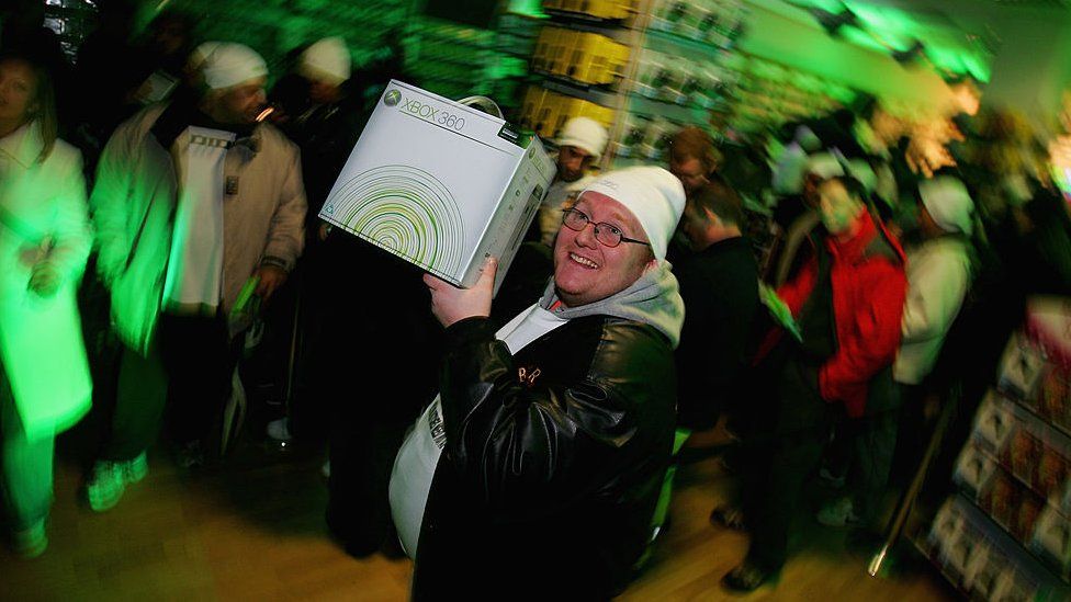 A gamer wearing a white beanie hat and black leather jacket with grey hoodie underneath proudly holds up the box for his new Xbox 360. The background of a busy games shop is visible but blurry behind him