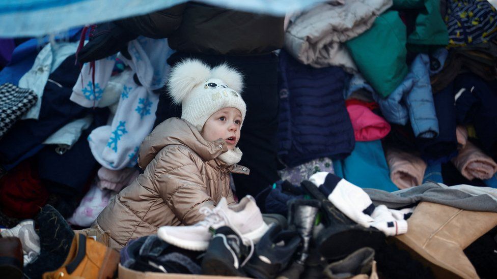 A small child walks around a pile of clothes at a makeshift camp