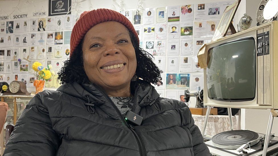 A woman in a hat sits in front of an exhibition of local Windrush generation people who have passed away.