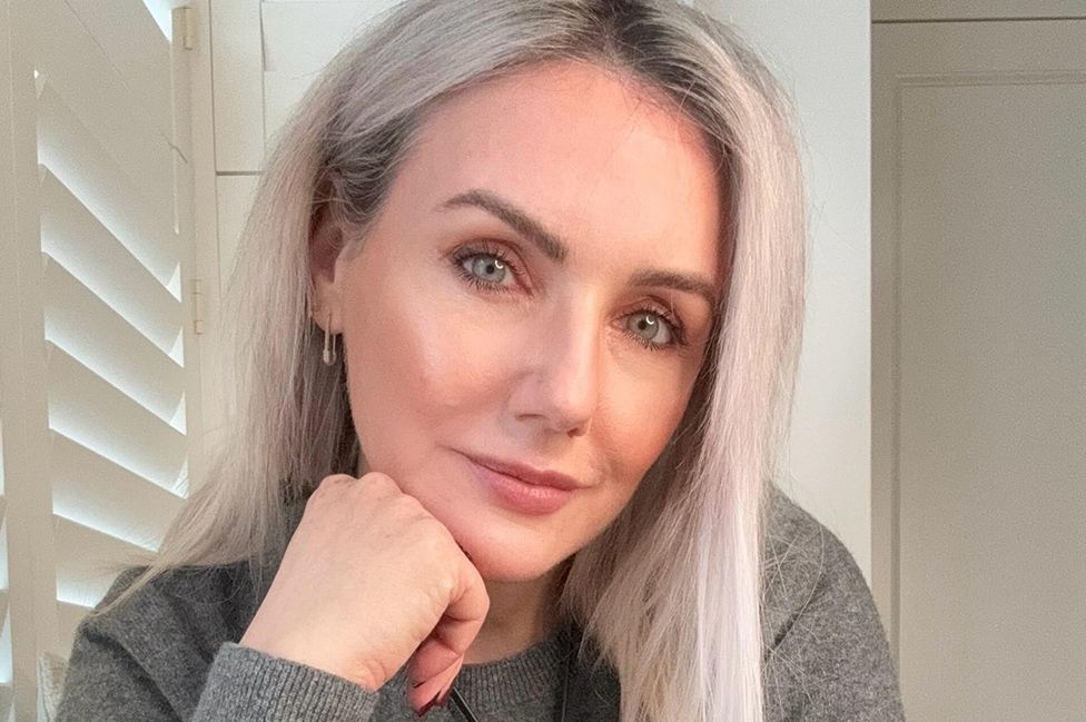 A woman sits near a window with wooden blinds, resting her chin on her hand. She's smiling slightly, her pale blue eyes are piercing. She has grey hair cut into a mid-length style and wears a grey sweater.
