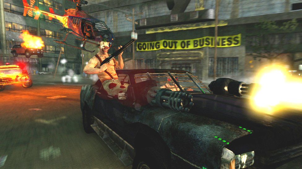 A chaotic screenshot from a video game shows a shirtless man in a clown mask sitting on the lip of a car door as he holds a large shotgun. The modified sports car he's a passenger in is fitted with two large gatling gun weapons - a bright yellow muzzle flash emanates from one. Behind him is a post-apocalyptic scene - we can see a building with a large, yellow "going out of business" banner across its front. A helicopter flies through the shot, a parked car appears to be on fire and a third, modified vehicle appears to be chasing the car carrying the man with the shotgun.