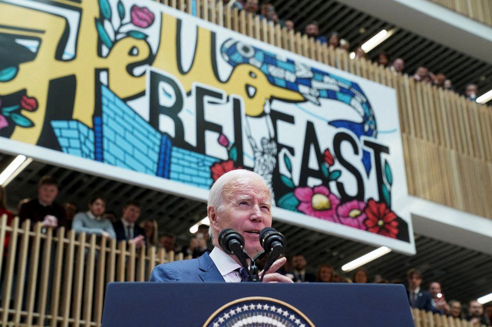 People watch as Joe Biden gives a speech beneath a large mural that reads: Hello Belfast