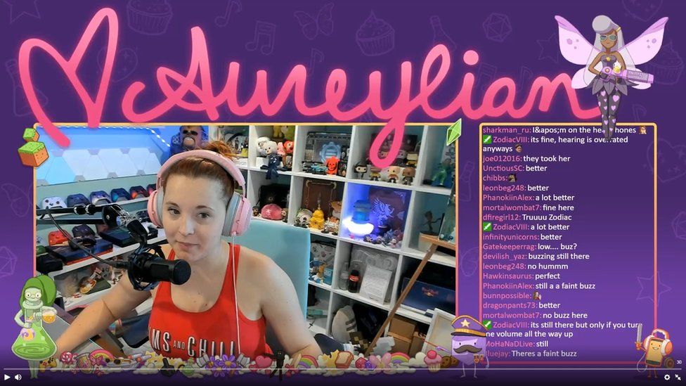 A screenshot of a Twitch stream. In the middle is the video stream featuring Aureylian, a young woman wearing a headset in front of a microphone. In the background are various gaming consoles and controllers, as well as figurines and models. The screenshot also shows a chat screen filled with various comments from her fans.