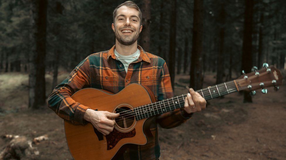 Sam Evans holding a guitar in a forest
