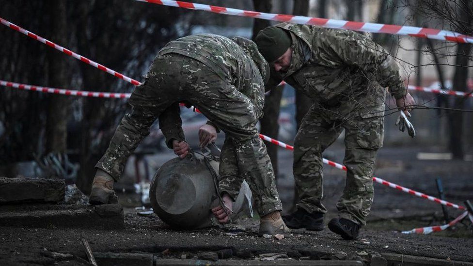 Two people in military fatigues lean over a large round metal item