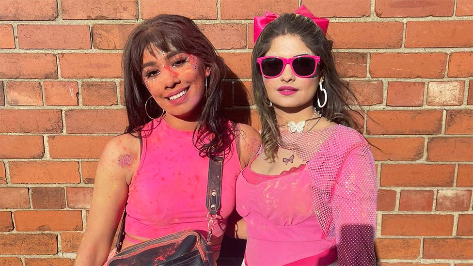 Shaz and Mahum - two female friends standing next to each other. Shaz on the left is smilng and has pink powdered colour on her face, with silver hoop earrings, wearing a pink vest top with a black bag strap hanging. Mahum on the right is wearing sunglasses with pink rims, white hoop earrings and a pink top, with a pink bow in her hair. The background is of a brick wall.