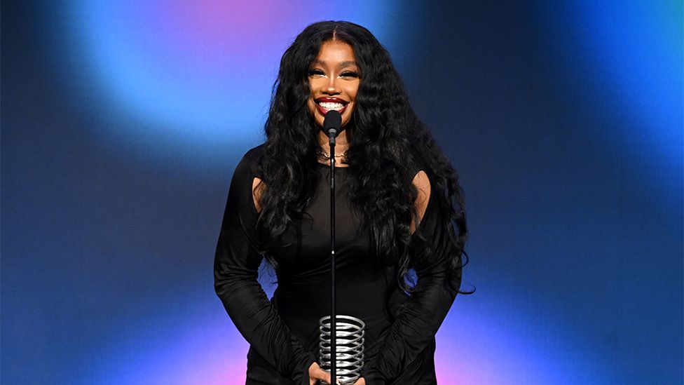 SZA, a woman wearing a black outfit, standing on stage behind a microphone, accepting an award. She is smiling. The background is dark and light blue and pink.