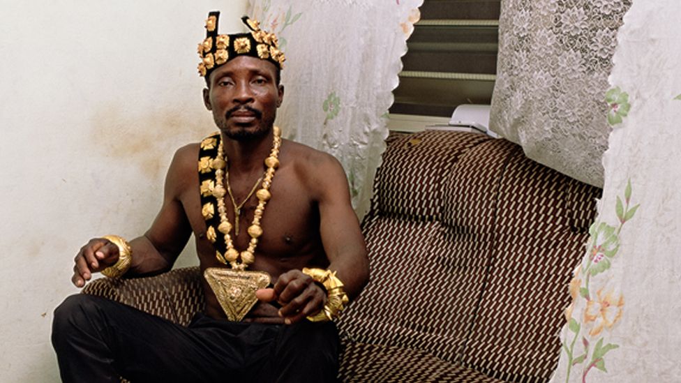 A photo by Deana Lawson of a shirtless man sitting on a sofa