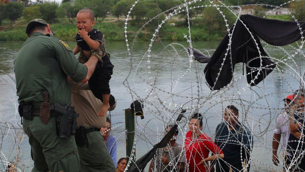 A Border Patrol agent carries one year-old Juan Diego ahead of his mother through the opening they made in the razor wire to allow migrants from Venezuela to enter after they crossed the Rio Grande in Eagle Pass, Texas