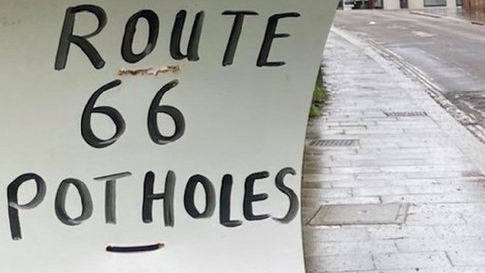 White sign with black lettering saying "Route 66 Potholes" on urban road