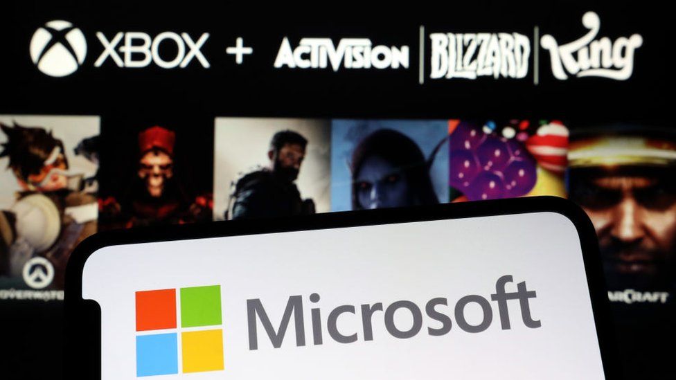 A smartphone shows the Microsoft logo of a red, green, yellow and blue square against the company's name on a blank screen. The background is blurry but outlines of characters from well-known Blizzard and Activision titles can be seen. Above them, the Xbox, Activision, Blizzard and King company logos are visible