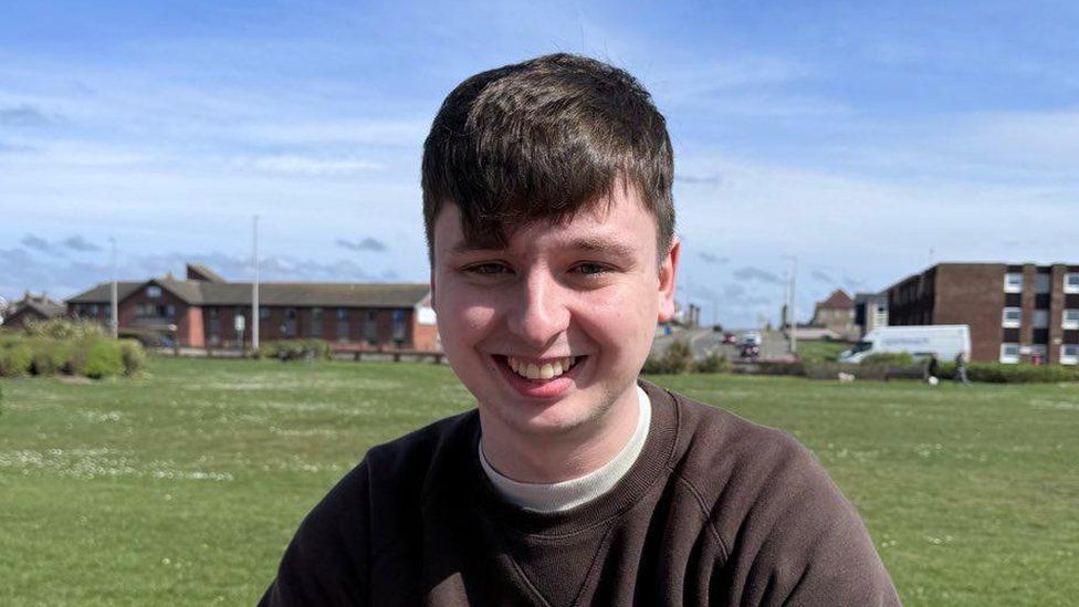 Ben Thomas, a white man in his twenties, with brown hair and a fringe smiles into the camera.