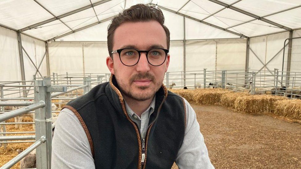 Jack Pishhorn with very short dark beard and glasses sitting on a straw bale in a marquee