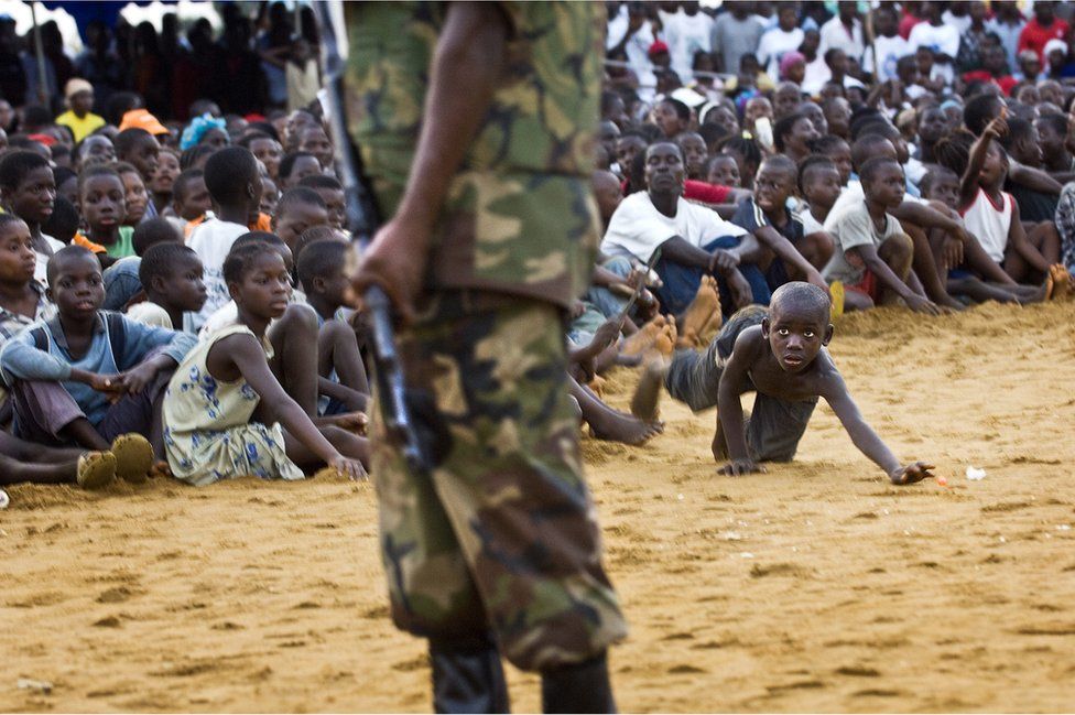 A boy from a crowd retrieves his ball from the shadow of armed security at the Liberia National Peace and Cultural Festival in Monrovia