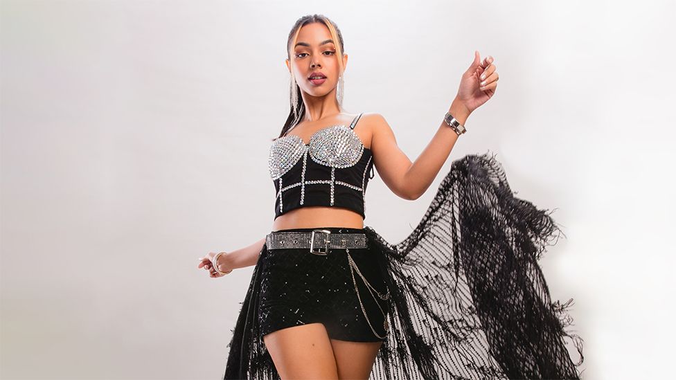Ri (Riya Duggal), a woman, is wearing a silver and black top and black shorts with a large bird like feather around her. She is looking into the camera and has silver earrings