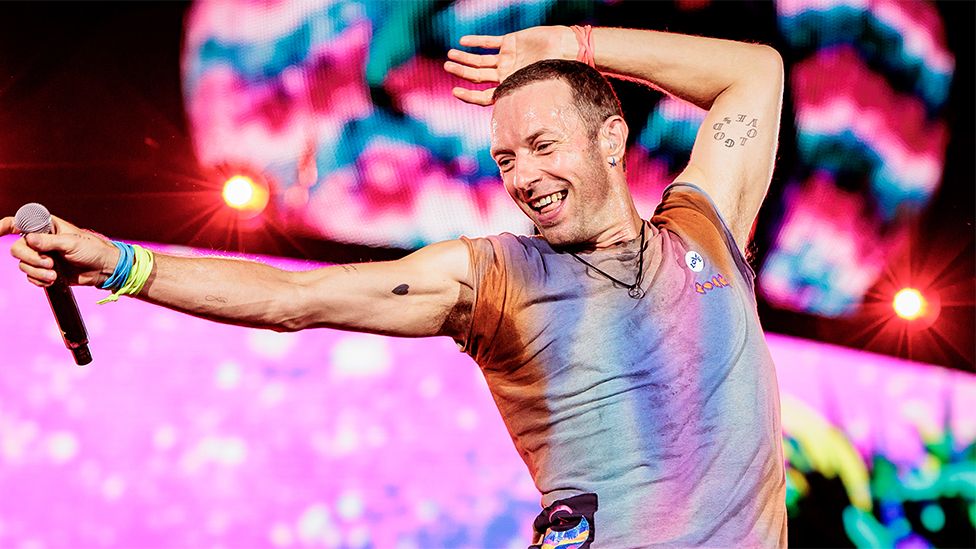 Chris Martin from Coldplay, a man wearing a colourful vest pointing with both hands to his right. He has a black microphone in his right hand and blue and green wrist bands. The background is flashing pink patterns with blue.