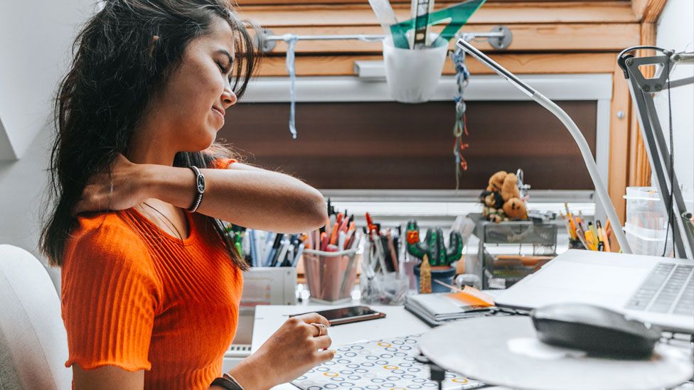 A woman rubs her shoulder whilst working at home