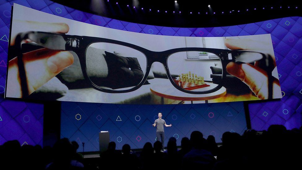 Mark Zuckerberg on stage with a large screen above him showing a pair of glasses.