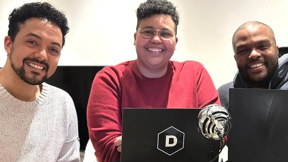 Two men and one woman smile while looking at the camera. They're sitting behind two laptops and a microphone, as well as a pile of dice