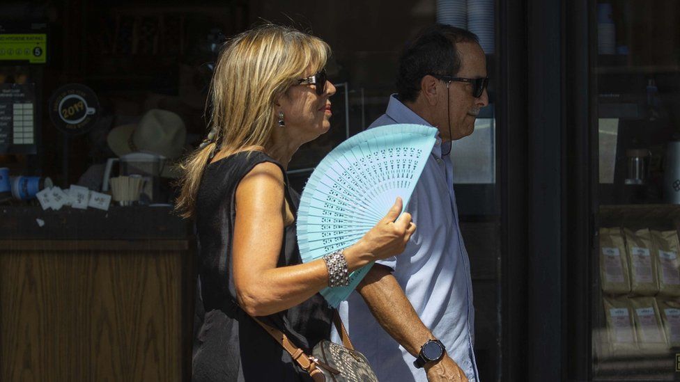 Woman walks down street using blue fan to cool herself, man walks beside her