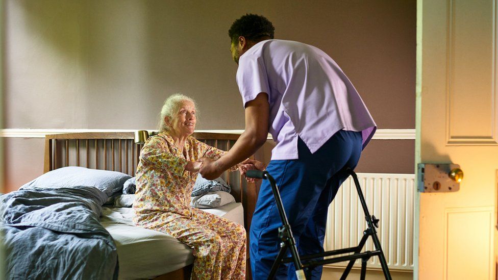 Young male care worker helping woman off bed with walking frame nearby - stock photo