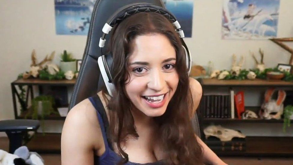 A photo of anita sat in a gaming chair with a headset on, she is smiling and laughing whilst looking into the camera with a bookshelf and posters in the background.