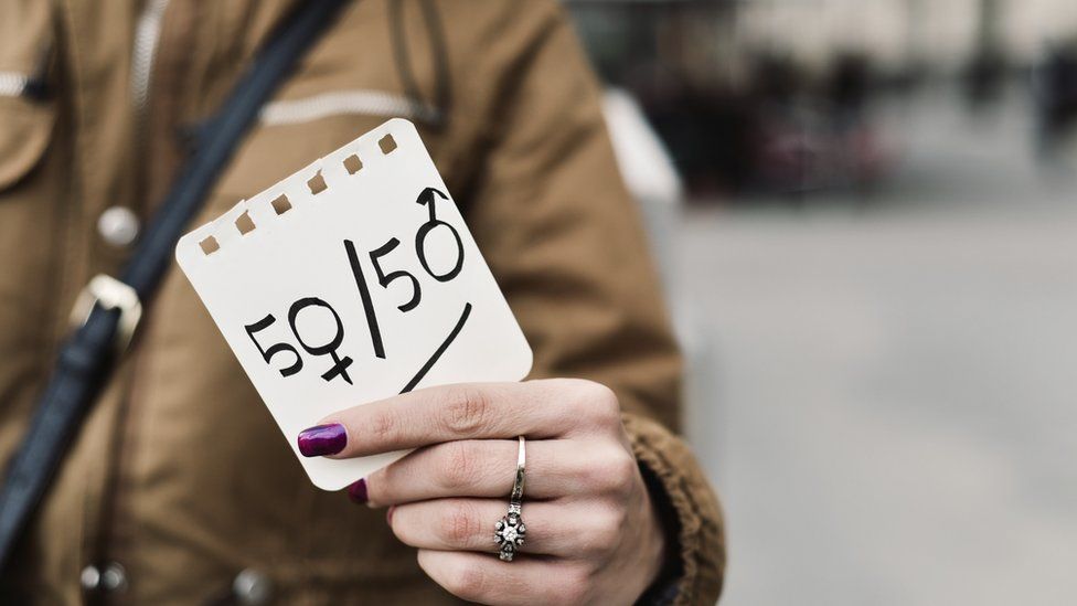 A woman in the street shows a paper with the text 50/50 written in it, with the zeros as the female and male gender symbols, depicting the gender parity concept.