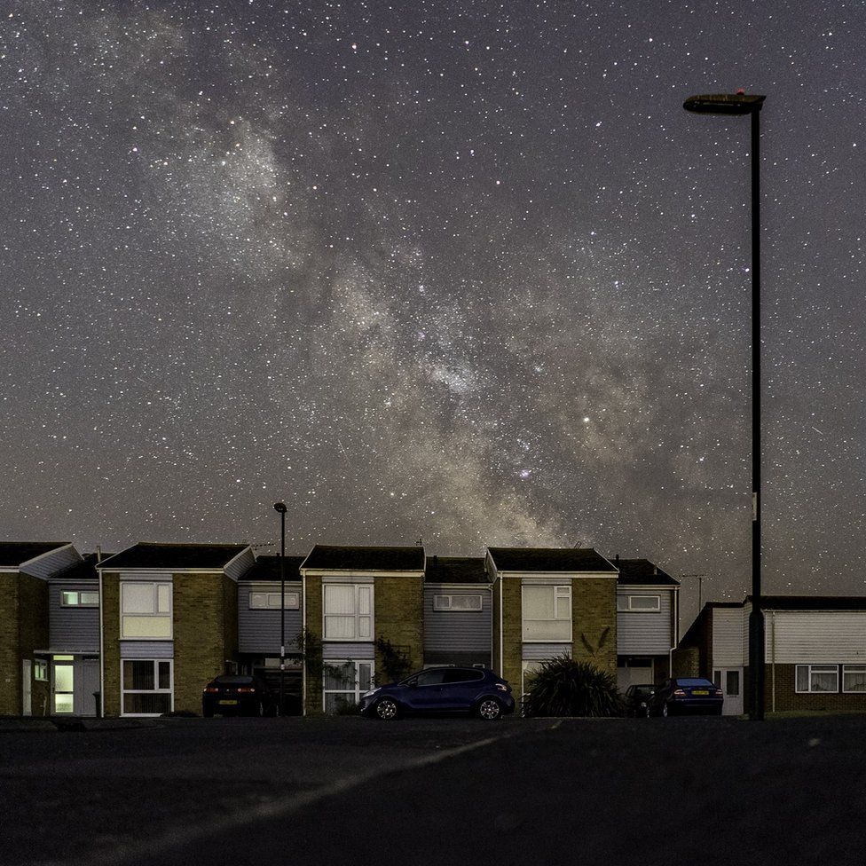 A silhouette of a lamppost reaches up from suburbia into the milky way