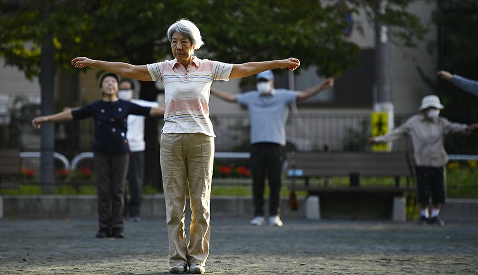 People exercising in a park in Tokyo in October 2022 - person in foreground with grey hair extends shoulders while standing up straight