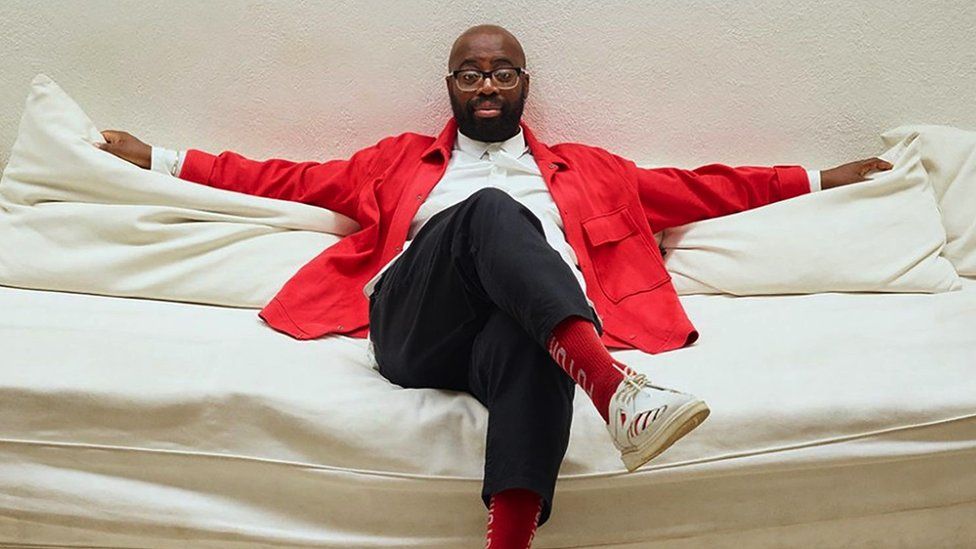 A man wearing a bright red jacket sits on a couch with his arms spread out