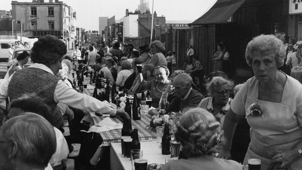People gathered around a long table set with bottles and food for a street party for the Queen's Jubilee in 1977